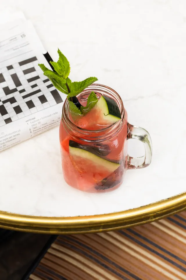 Cocktail met munt en fruit in een glas op een marmeren tafeltje naast een krant en een pen.