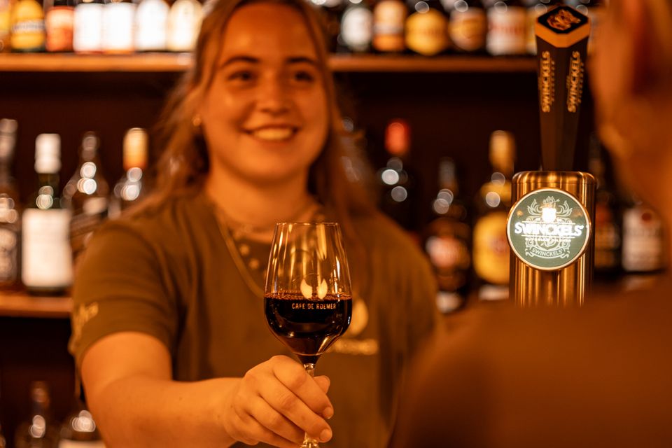 Lachende medewerker achter de bar schenkt een glas rode wijn, met flessen en tapkranen erachter.
