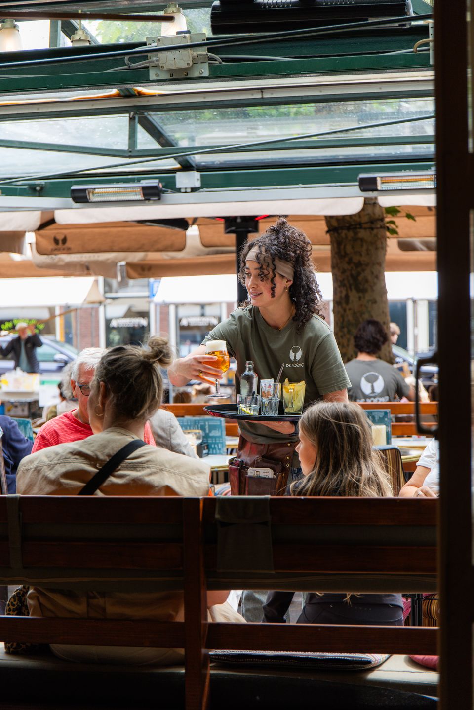 Medewerker brengt drankjes naar gasten in de serre, met het terras en de Botermarkt op de achtergrond.