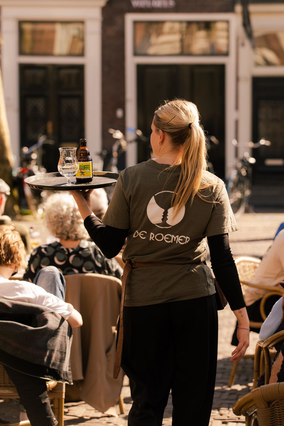 Medewerker met een dienblad vol drankjes loopt tussen de gasten op het zonnige terras aan de Botermarkt.