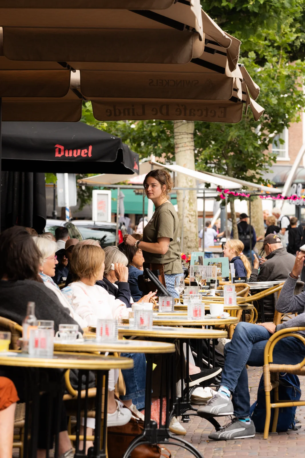 Medewerker in een groen Roemer-shirt serveert gasten op het zonnige terras onder een bruine parasol.